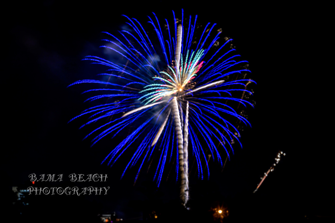 Blue and multicolored fireworks opening against a black Alabama Gulf Coast night sky.