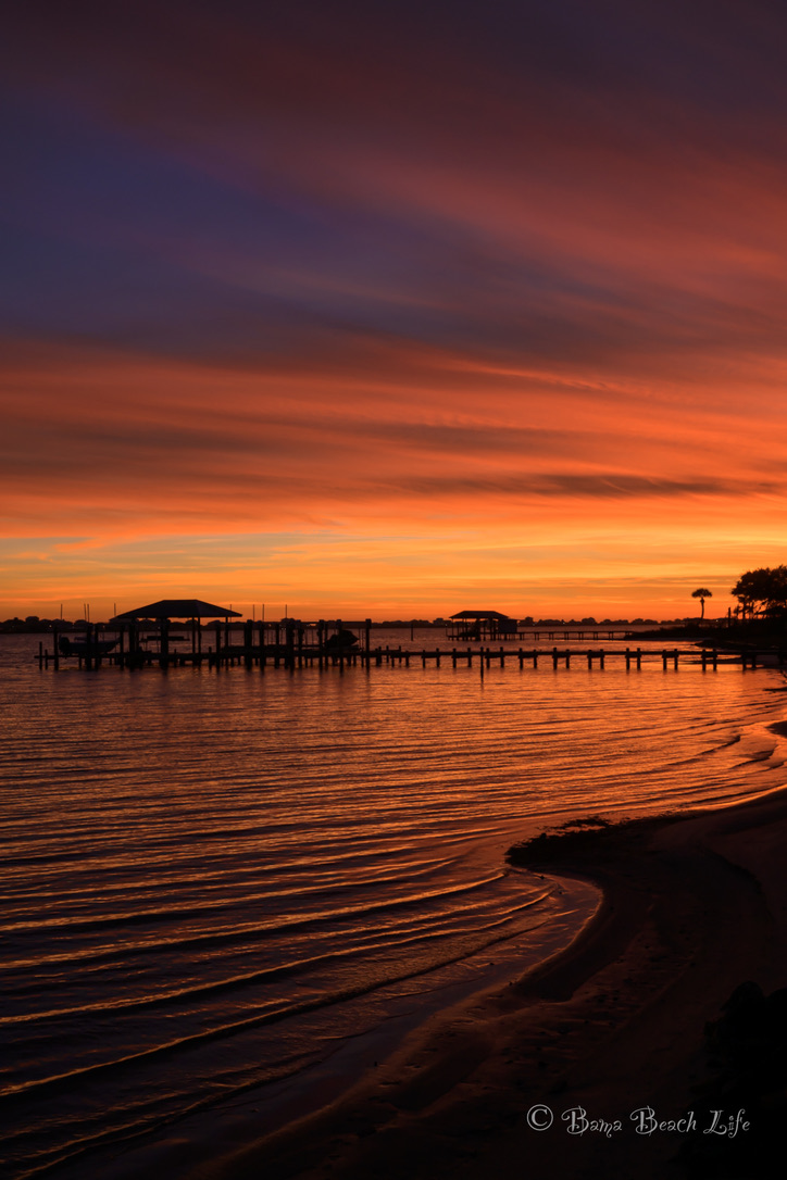 Fiery orange sunset illuminating docks, rippled water, and shoreline at Little Lagoon in Gulf Shores.
