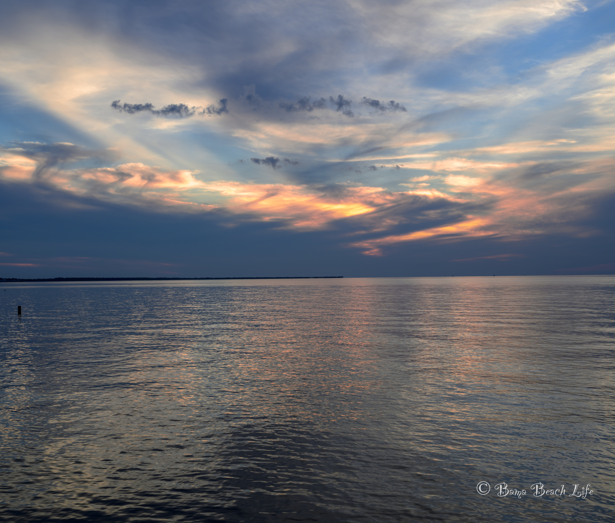 Layered blue, coral, and gold sunset clouds reflected across calm Gulf of Mexico water.