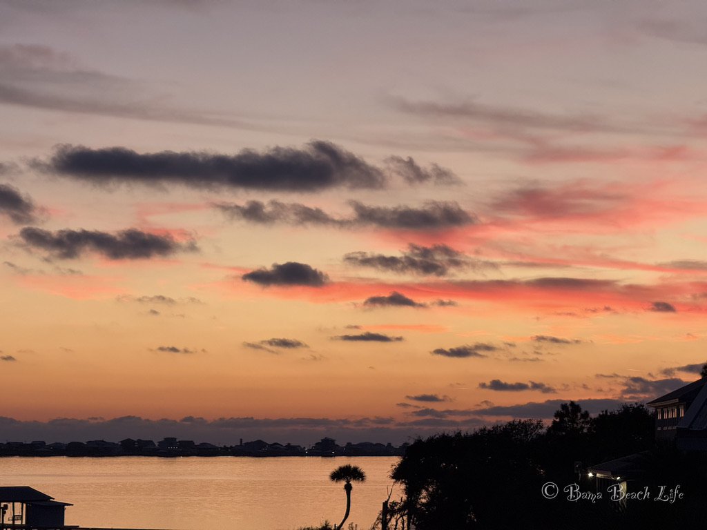 Pastel coral clouds over lagoon water and a silhouetted coastal shoreline in Gulf Shores, Alabama.
