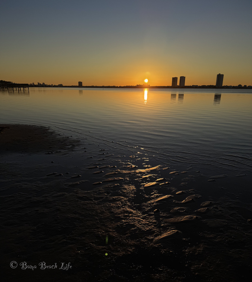 Rising sun reflected in the calm water and wet sand of Little Lagoon in Gulf Shores.