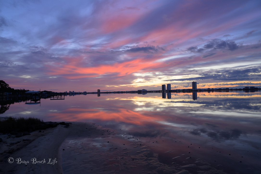 Pink and violet sunset clouds mirrored across still Little Lagoon water in Gulf Shores, Alabama.